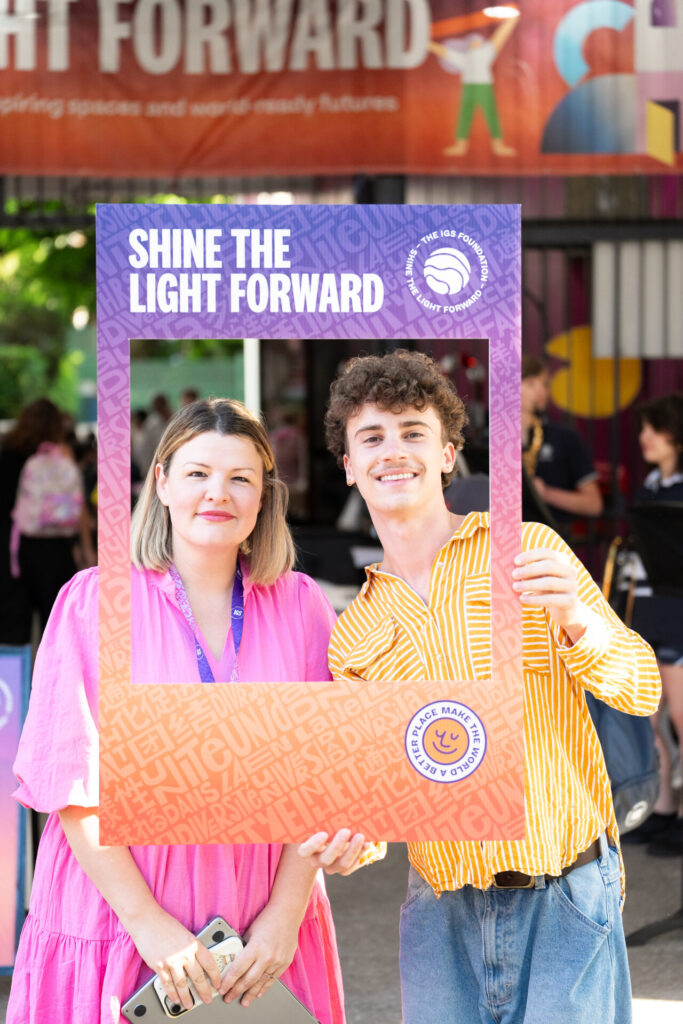 Two people smile and pose outdoors, holding a colourful Shine the Light Forward photo frame. The woman wears a pink dress, and the man wears a yellow striped shirt. A festival banner is visible in the background.