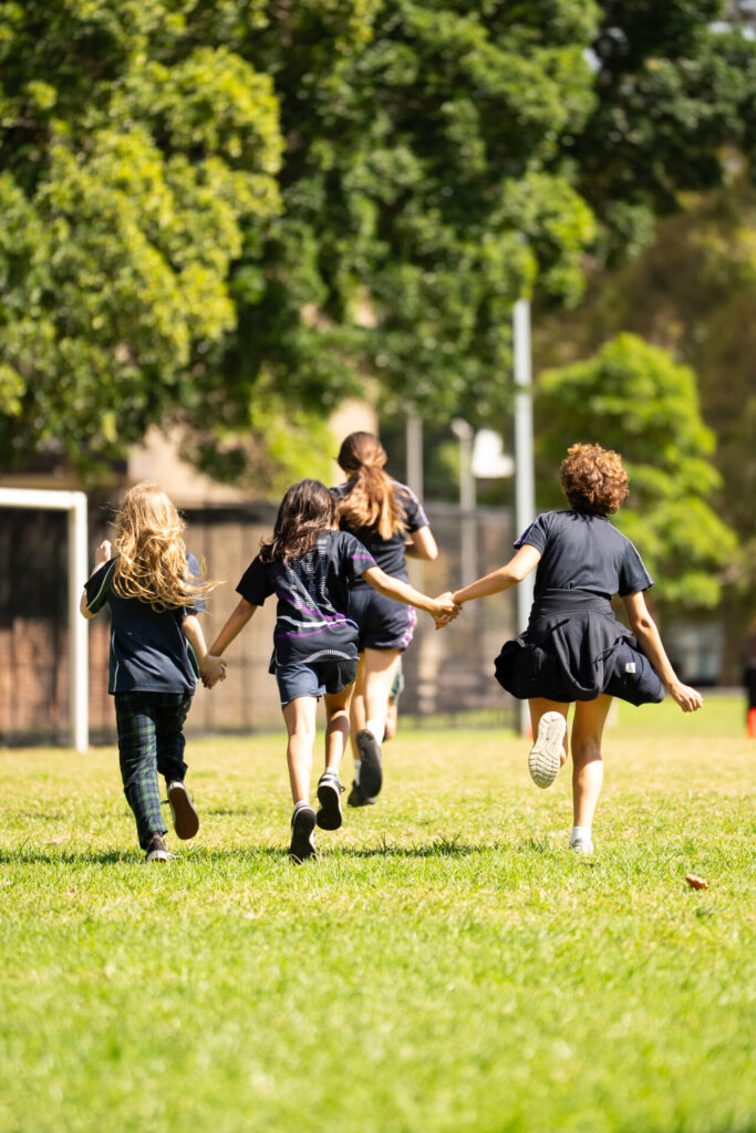 Four children run hand-in-hand across a grassy field towards trees, seen from behind on a sunny day. They appear joyful and energetic, enjoying outdoor play.