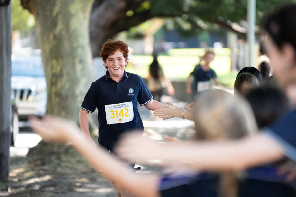 A young runner with curly red hair, wearing a dark shirt and race number 3142, smiles whilst high-fiving supporters along a sunlit, tree-lined path during a race.
