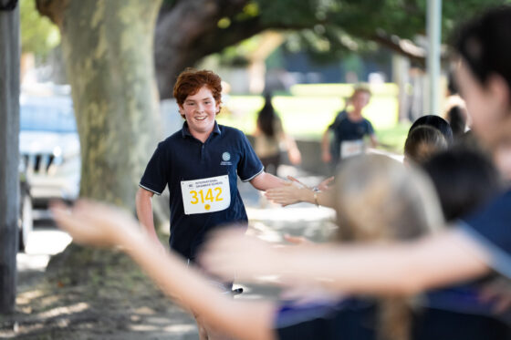 A young runner with curly red hair, wearing a dark shirt and race number 3142, smiles whilst high-fiving supporters along a sunlit, tree-lined path during a race.