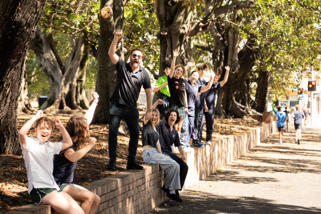 A group of people sit and stand on a low brick wall under large trees, smiling and raising their arms in excitement. Sunlight filters through the leaves and the street is visible in the background.