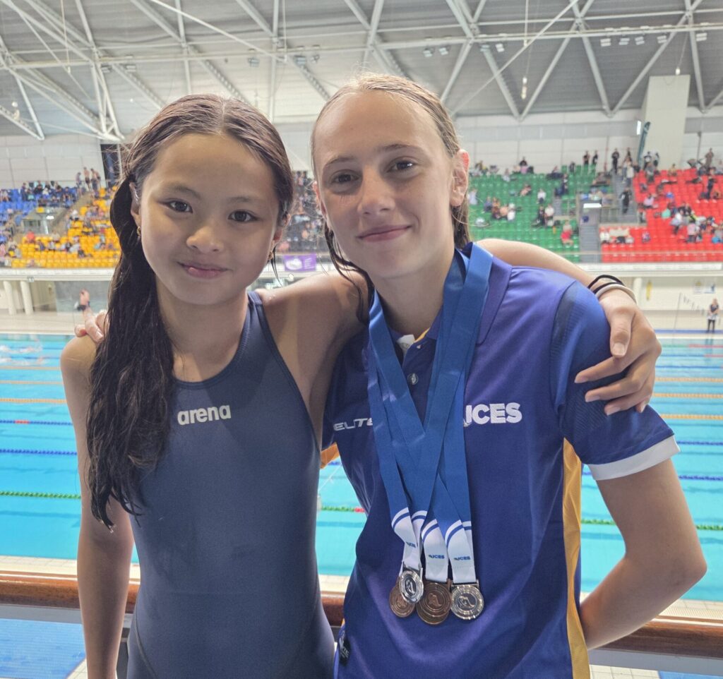 Two young swimmers stand side by side at an indoor pool, embodying sportsmanship. One wears a dark swimming costume; the other, in a blue shirt with medals around her neck, has her arm around her team-mate. Spectators fill the colourful seats behind them.