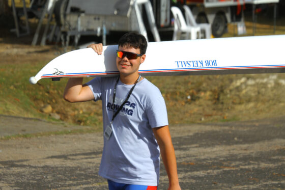 A person wearing sunglasses and a Rowing T-shirt demonstrates true sportsmanship as they carry a white rowing boat labelled BOB RELFALL on their shoulder outdoors, with chairs and equipment in the background.