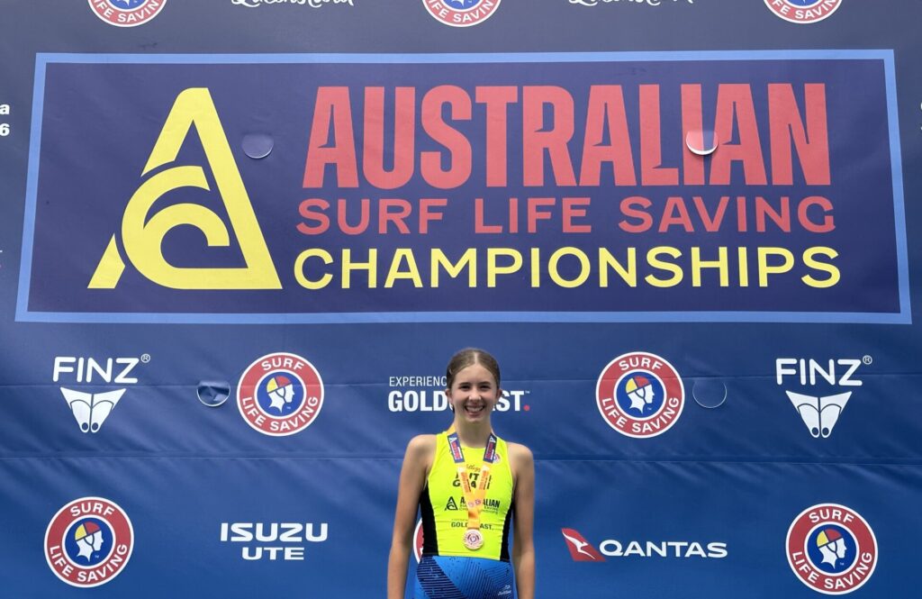 A young athlete, embodying the spirit of sport, smiles in front of a large banner that reads Australian Surf Life Saving Championships, with sponsor logos and medals round her neck.