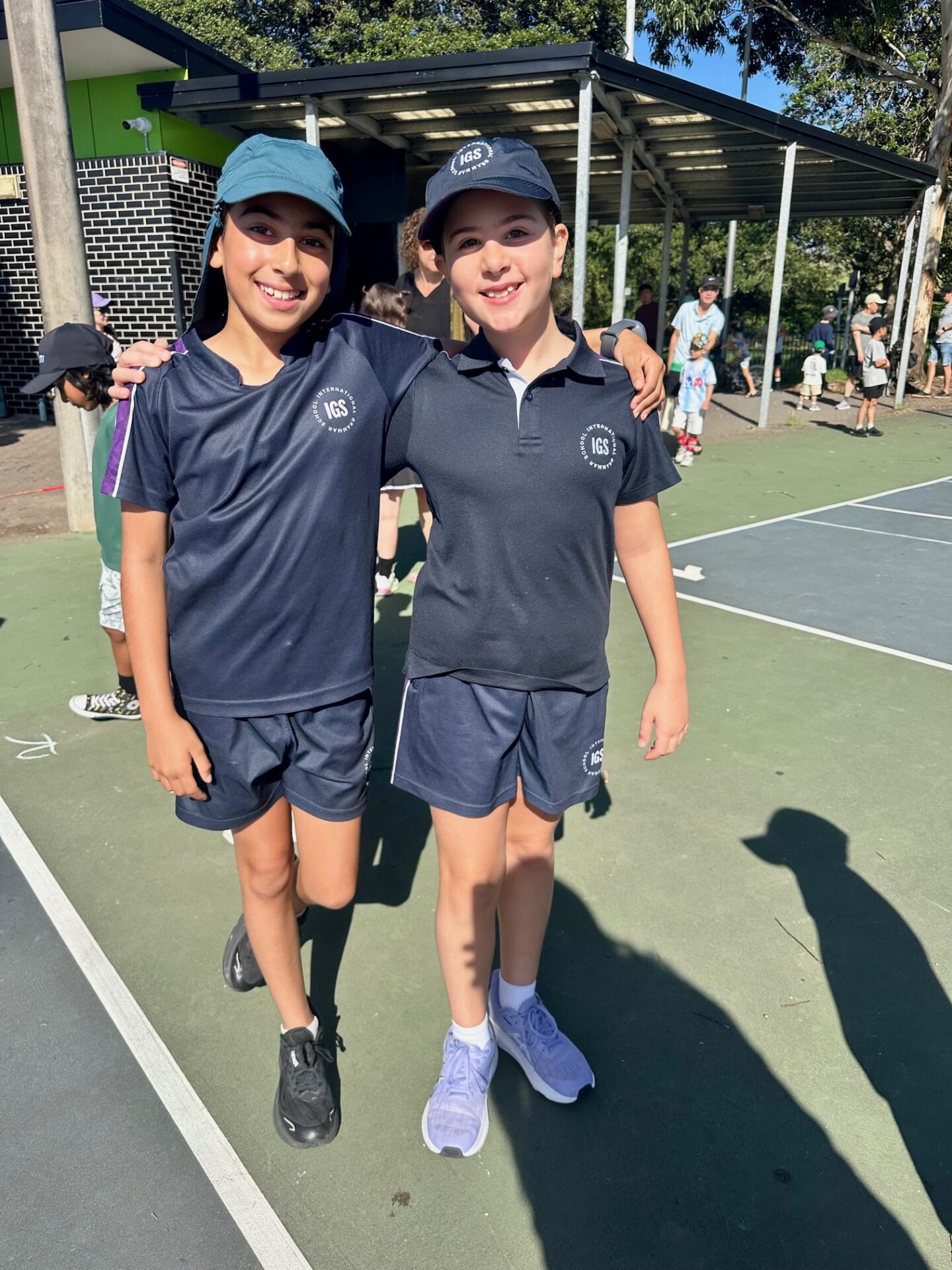 Two children in navy sports kits and caps stand arm-in-arm on a sunny outdoor court, smiling at the camera. Their spirit shines as they enjoy the sport, with other children visible in the background near a covered area.