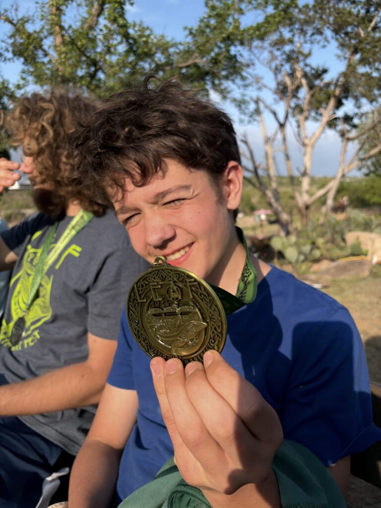 A smiling teenage boy named Kasper in a blue shirt holds up a gold medal outdoors, with trees and another person in the background. Sunlight highlights his face and the medal as he proudly shows off his medals collection.