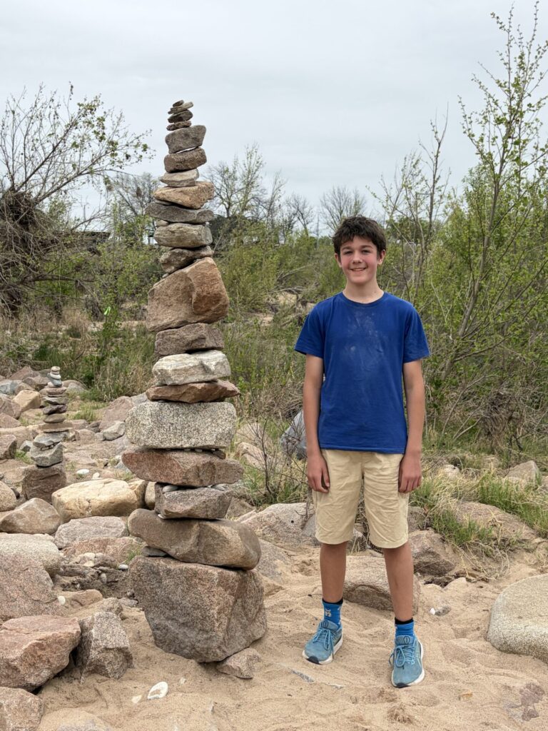 A smiling boy in a blue shirt and khaki shorts stands on sandy ground beside a tall Kasper Stacks rock cairn. Bushes and rocks fill the outdoor background under a cloudy sky.