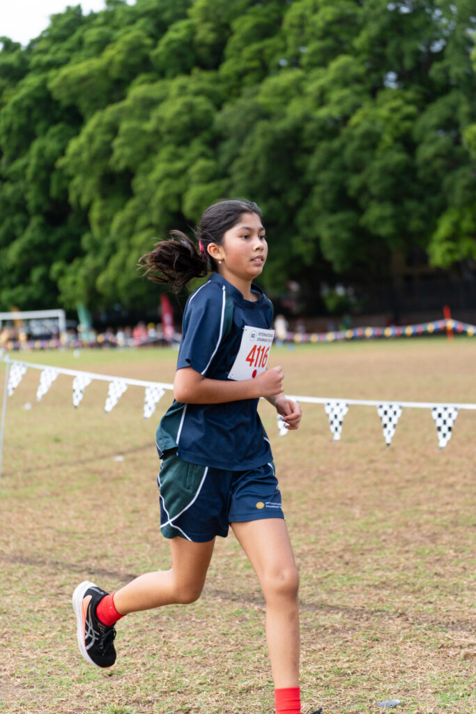 A girl in sports kit and a race number runs on a grassy field during a race, with trees and a chequered flag banner in the background.