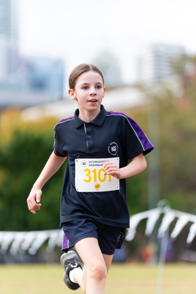 A young girl in a navy sports kit with the number 310 runs outdoors during a race, with buildings and trees blurred in the background.