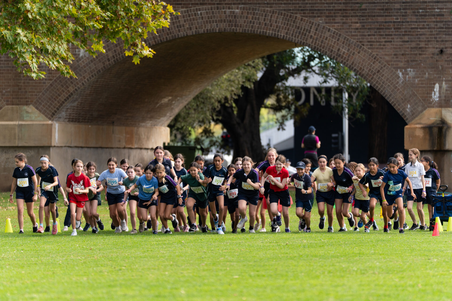 A large group of children wearing sports clothes start running a race on grass under a brick archway, with trees and cones visible in the background.