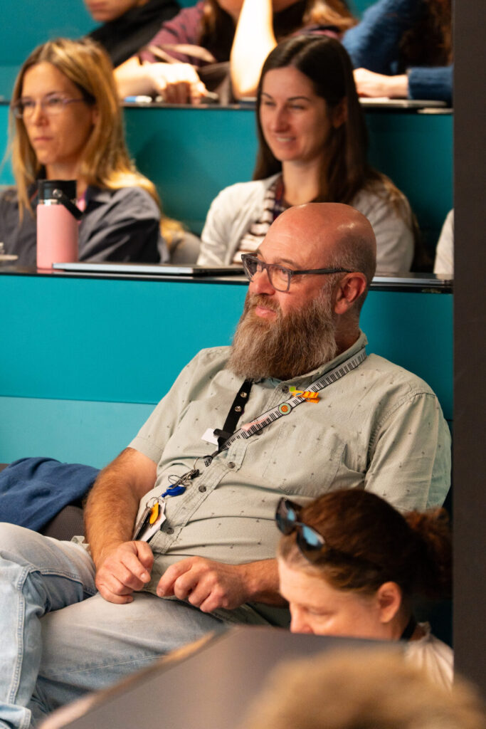 A bearded man with glasses sits and smiles in a lecture theatre among other attendees, all listening attentively to a discussion on Truth to Power. The background features teal walls and people with notebooks and water bottles.
