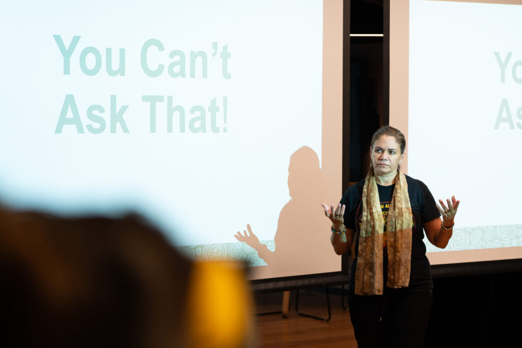 A woman stands in front of a screen displaying the words You Can't Ask That! She gestures with her hands, confidently addressing the audience about truth and power during her presentation.