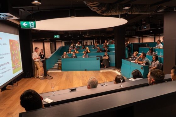 A group of people sit in a modern, tiered lecture theatre watching a presenter discuss the state of play in digital gaming. The engaged audience listens closely, surrounded by teal seating and wood flooring.
