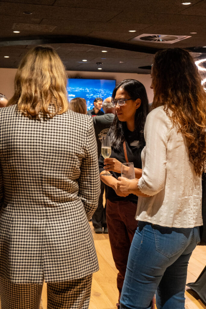 Three women stand in a group at an indoor social event, talking and holding drinks. The lively setting features other guests and a large screen in the background displaying highlights from the latest State of Play digital gaming showcase.