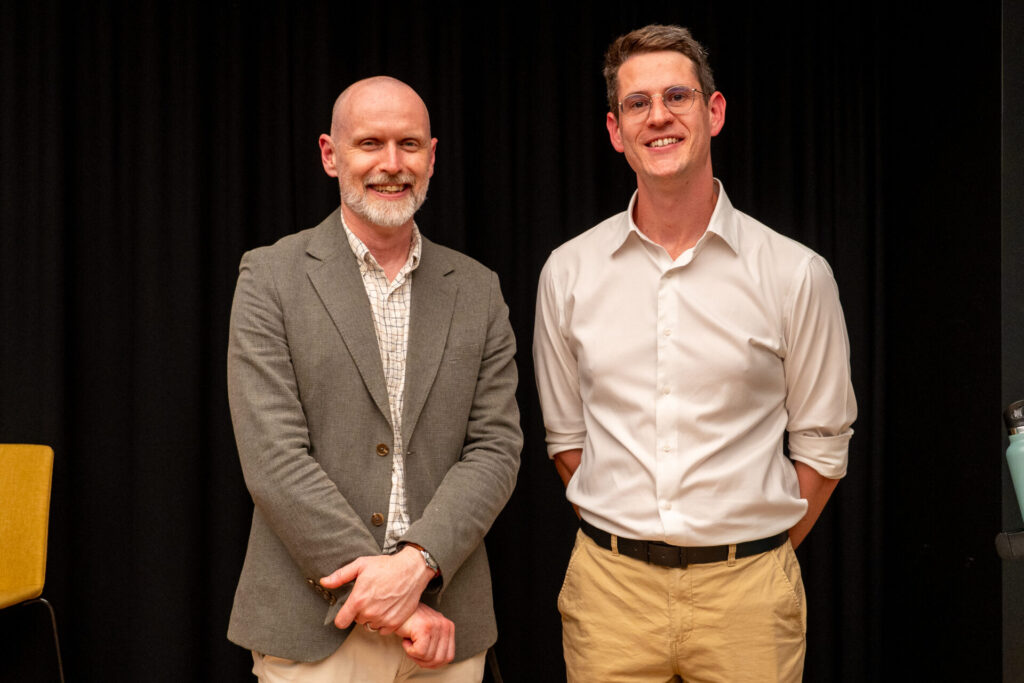 Two men stand side by side, smiling at the camera. One wears a grey blazer and checkered shirt, while the other sports glasses, a white shirt with rolled sleeves, and khaki trousers. They appear at a State of Play digital gaming event with a black background behind them.
