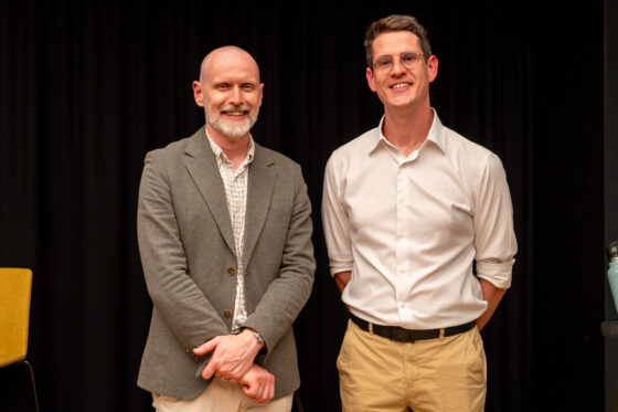 Two men stand side by side, smiling at the camera. One wears a grey blazer and checkered shirt, while the other sports glasses, a white shirt with rolled sleeves, and khaki trousers. They appear at a State of Play digital gaming event with a black background behind them.