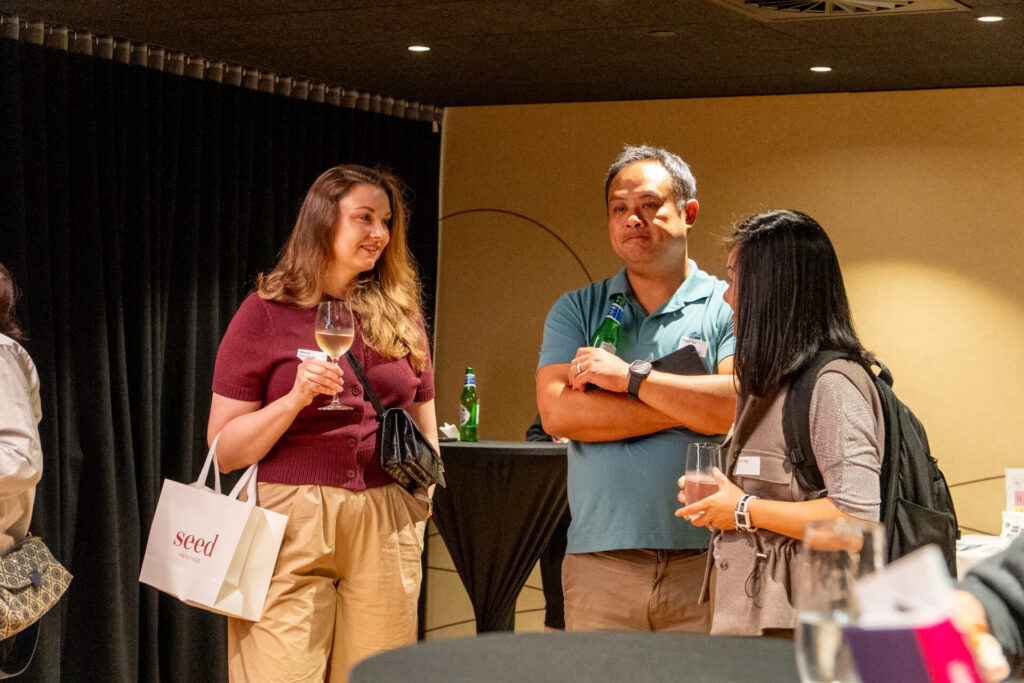 Three people stand and chat at an indoor event. One woman holds a drink and a gift bag, while a man with arms crossed holds a bottle. Nearby, "State of Play" banners hint at a digital gaming theme as they appear relaxed and engaged in conversation.