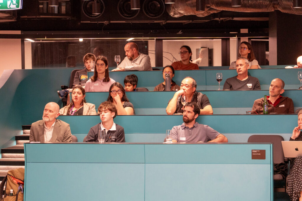 A diverse group of people sit in tiered teal lecture theatre seats, attentively listening to a State of Play presentation on digital gaming. Some have drinks in front of them, and a few appear to be taking notes. The atmosphere is focused and engaged.