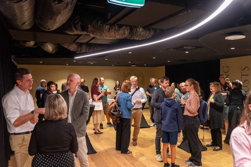 A group of people stand and talk in a modern, well-lit room with wooden floors. Some discuss the State of Play in digital gaming, holding drinks or papers, whilst others mingle in small groups. Large ducting is visible on the ceiling.