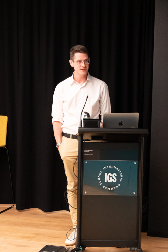 A man in a white shirt and beige trousers stands at a lectern with a laptop, microphone, and IGS Internationale Gymnasium sign, discussing the State of Play in Digital Gaming in front of a black curtain.
