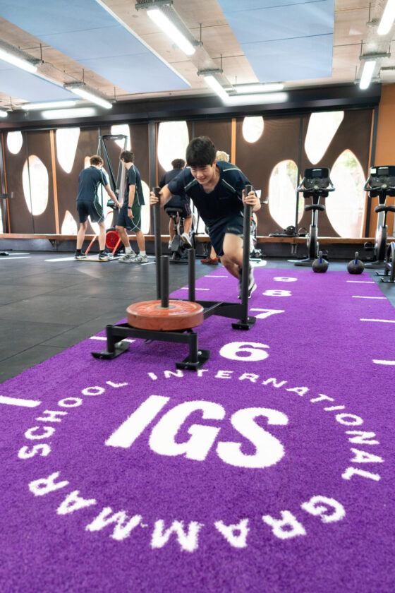 A student pushes a weighted sled on a purple mat labelled IGS GRAMMAR SCHOOL INTERNATIONAL in a gymnasium, building joy as other students exercise energetically in the background.