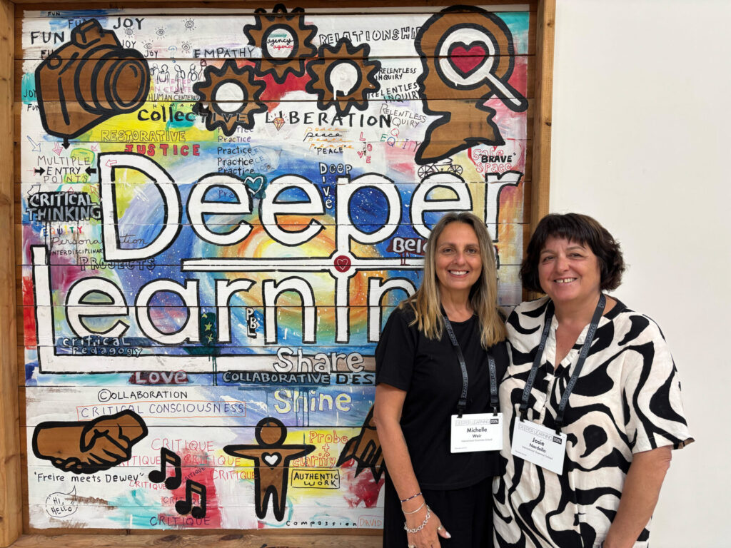 Two women smile in front of a vibrant mural reading Deeper Learning at the Deeper Learning Conference, surrounded by doodles and symbols of education, creativity, and collaboration. Both wear name badges on lanyards.