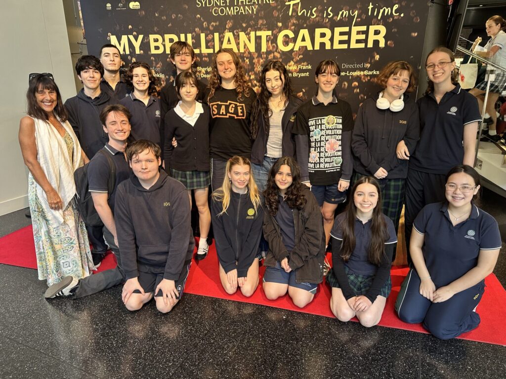 A group of students and an adult pose and smile on a red carpet in front of a My Brilliant Career Sydney Theatre Company backdrop, celebrating theatre education. Most are in school uniforms; the group looks happy and excited.
