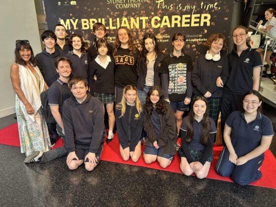 A group of students and an adult pose and smile on a red carpet in front of a My Brilliant Career Sydney Theatre Company backdrop, celebrating theatre education. Most are in school uniforms; the group looks happy and excited.