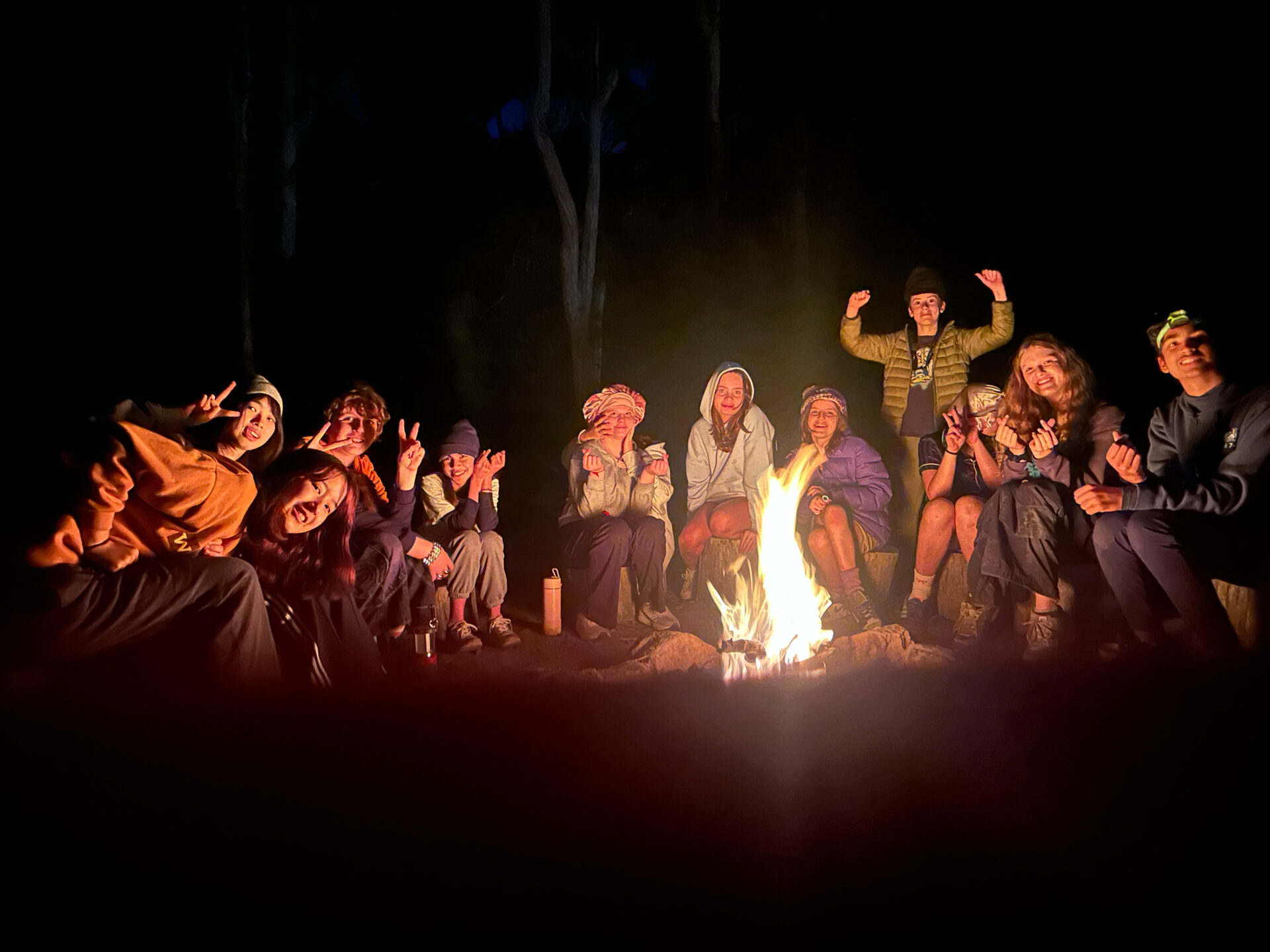A group of people sit in a circle around a campfire at night, smiling and posing for the camera. Enjoying experiential learning, they flash peace signs and wear warm clothing as the firelight illuminates their faces against the dark background.