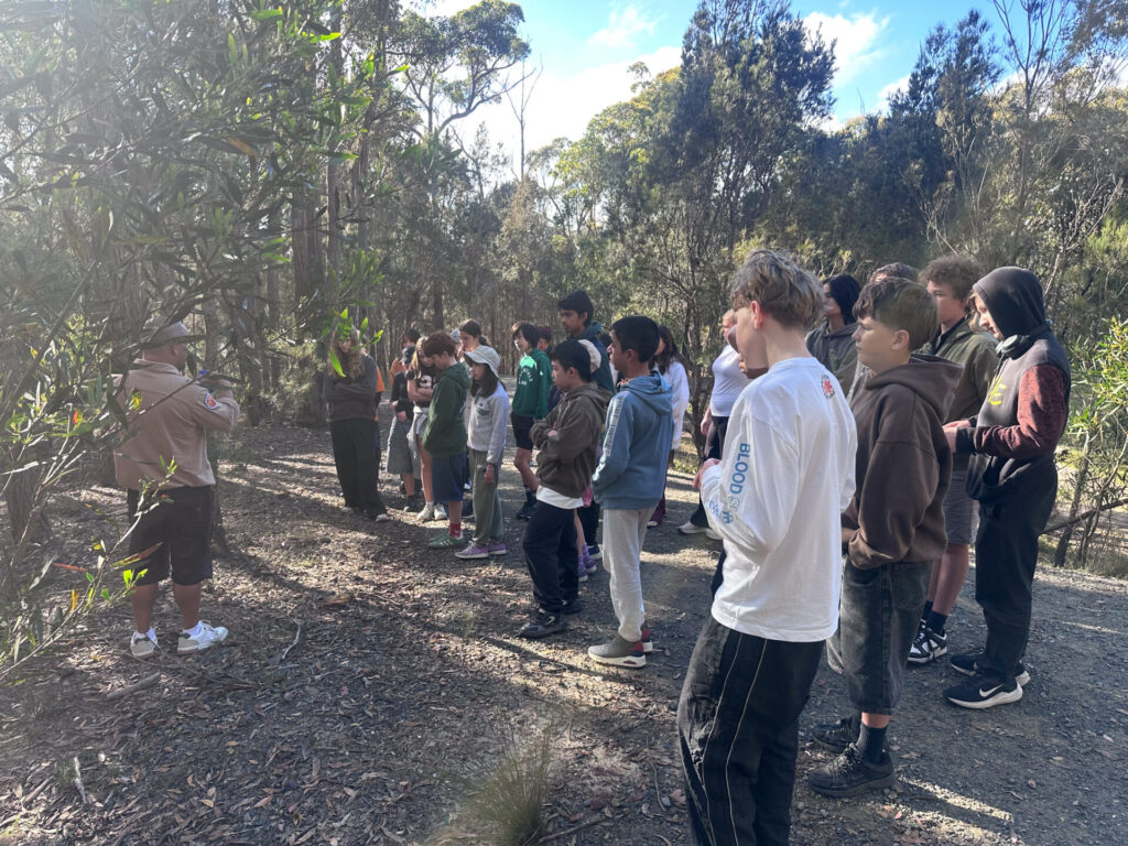 A group of students stands on a forest path, engaged in outdoor education as they listen to a guide. Dressed casually, they are surrounded by trees and natural vegetation under a partly cloudy sky, embracing experiential learning in nature.