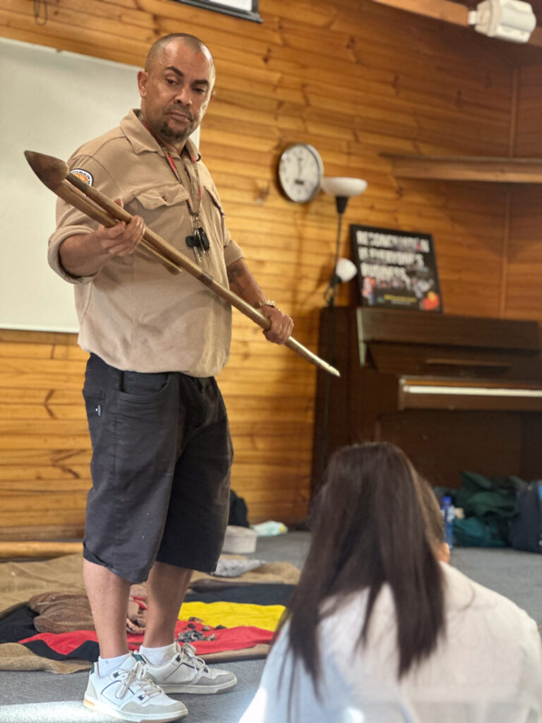 A man stands indoors holding a wooden spear and speaking to a seated person. Dressed in a beige shirt and black shorts, he appears to discuss bush activities. The room features wooden walls, a clock, a whiteboard, and a piano in the background.