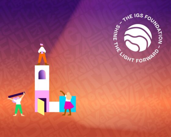 Three stylised figures build and stand on a tower, Building Joy as they go, with the words SHINE THE LIGHT FORWARD – THE IGS FOUNDATION encircling a wave-like logo on a purple and orange background with faint, scattered text.