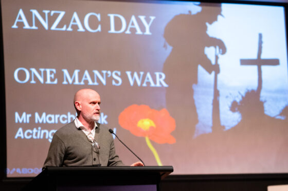 A man speaks at a lectern in front of a screen displaying ANZAC DAY: ONE MAN'S WAR with a silhouette of a soldier, a cross, and a red poppy in the background.
