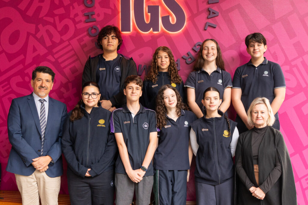 A group of eight Middle School Leaders in uniforms and two adults, one man in a suit and one woman in black, stand smiling before a pink wall with large text and the letters IGS above them, showcasing 2026 Middle School student leadership.