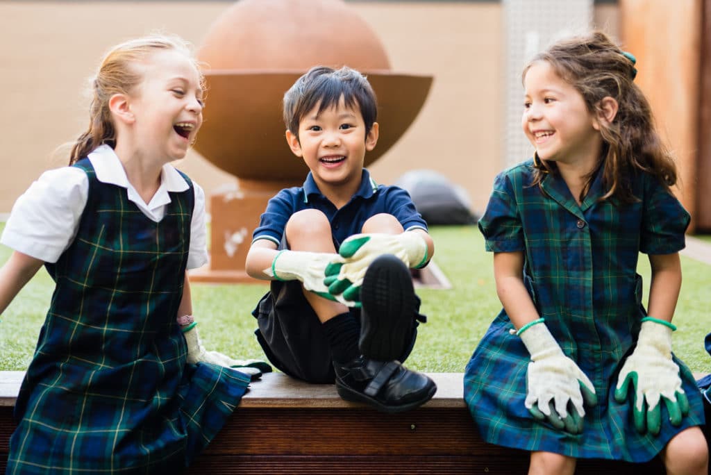 Three young children sit outdoors on a low wooden wall, smiling and laughing at their OSHC session. Two girls wear green checked uniforms and gardening gloves; a boy in the middle wears a navy shirt and gloves. A large garden ornament is in the background.