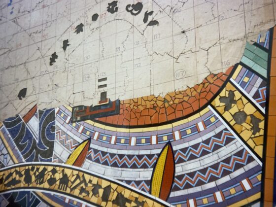 A colourful, geometric mural features abstract shapes and patterns with sections resembling maps and waves. The top portion appears weathered, with areas of missing material, while the lower section is vibrant and intact.