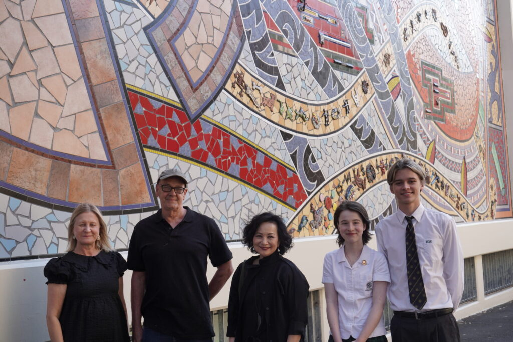 Five people stand in front of a colourful mosaic mural on an exterior wall, featuring abstract patterns, small figures, and a red bus. Three adults and two pupils are smiling at the camera.
