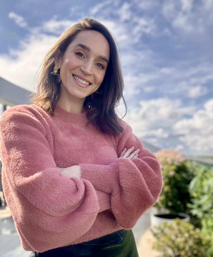 A woman with dark hair wearing a pink fluffy jumper stands outdoors with her arms folded, smiling at the camera. The background features greenery and a partly cloudy sky.
