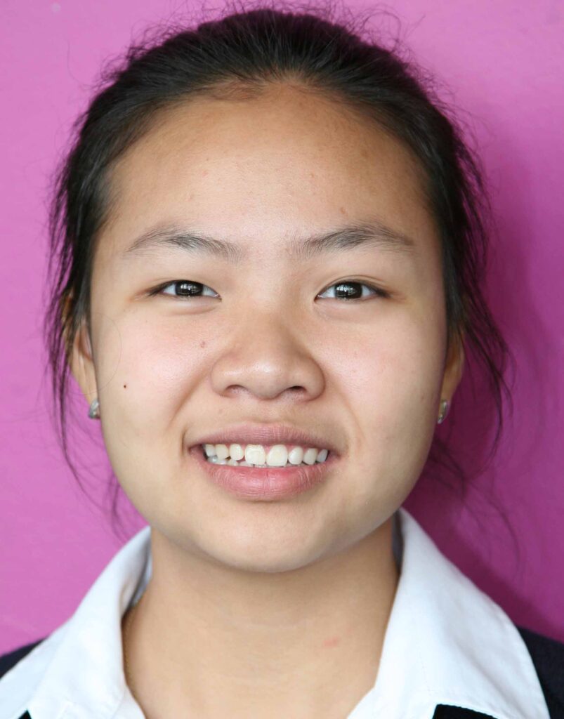 A young person with long dark hair pulled back, wearing a white collared shirt and a dark jumper, smiles in front of a plain pink background.