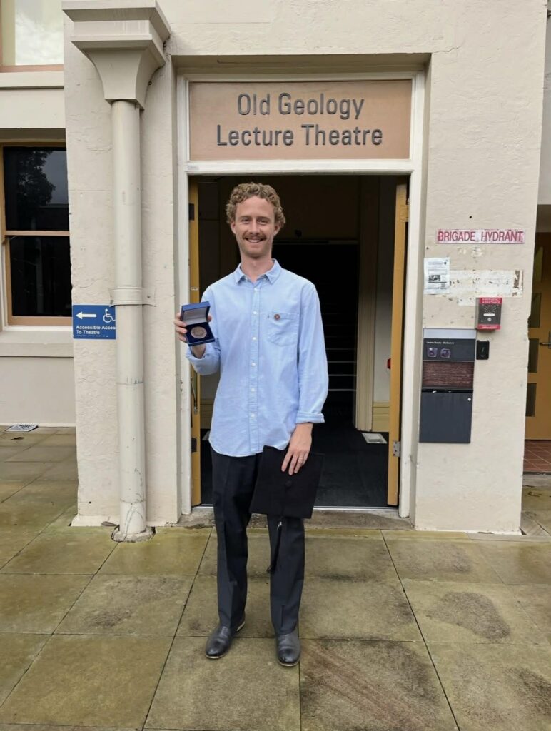 A young man with curly hair, wearing a light blue shirt and dark trousers, stands smiling in front of the Old Geology Lecture Theatre, holding a medal and a folder.