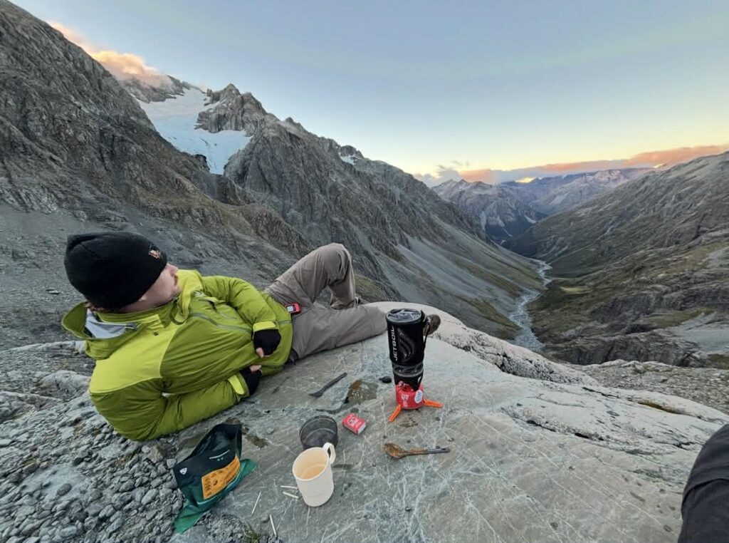 A person in a green jacket and black beanie relaxes on a rocky slope with camping kit, a hot drink, and a cooker, overlooking a dramatic mountain valley and distant glacier at sunset.