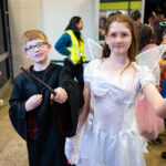 Two children in magical costumes stand indoors; the boy wears glasses, a black robe, and holds a wand, while the girl sports fairy wings, a white dress, and spectacles. Other people are visible in the background at the Primary Disco.