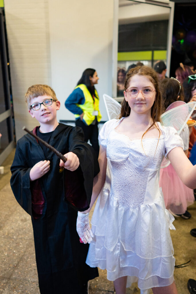 Two children in magical costumes stand indoors; the boy wears glasses, a black robe, and holds a wand, while the girl sports fairy wings, a white dress, and spectacles. Other people are visible in the background at the Primary Disco.