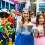 Three children in colourful primary costumes, including a panda hat, fairy wings, and a tiara, smile and eat hot dogs at a magical indoor party with disco lights and decorations sparkling in the background.