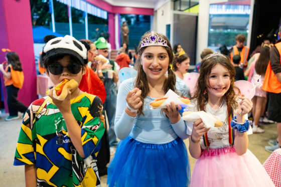 Three children in colourful primary costumes, including a panda hat, fairy wings, and a tiara, smile and eat hot dogs at a magical indoor party with disco lights and decorations sparkling in the background.