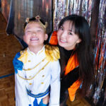 A smiling child in a magical prince costume and gold crown stands next to a woman with long dark hair in an orange waistcoat. They are posing happily at a primary school disco event, with shiny tinsel curtains sparkling behind them.