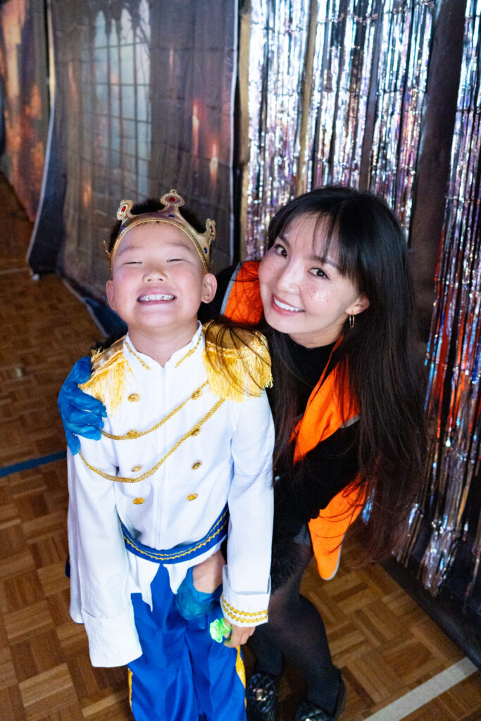 A smiling child in a magical prince costume and gold crown stands next to a woman with long dark hair in an orange waistcoat. They are posing happily at a primary school disco event, with shiny tinsel curtains sparkling behind them.