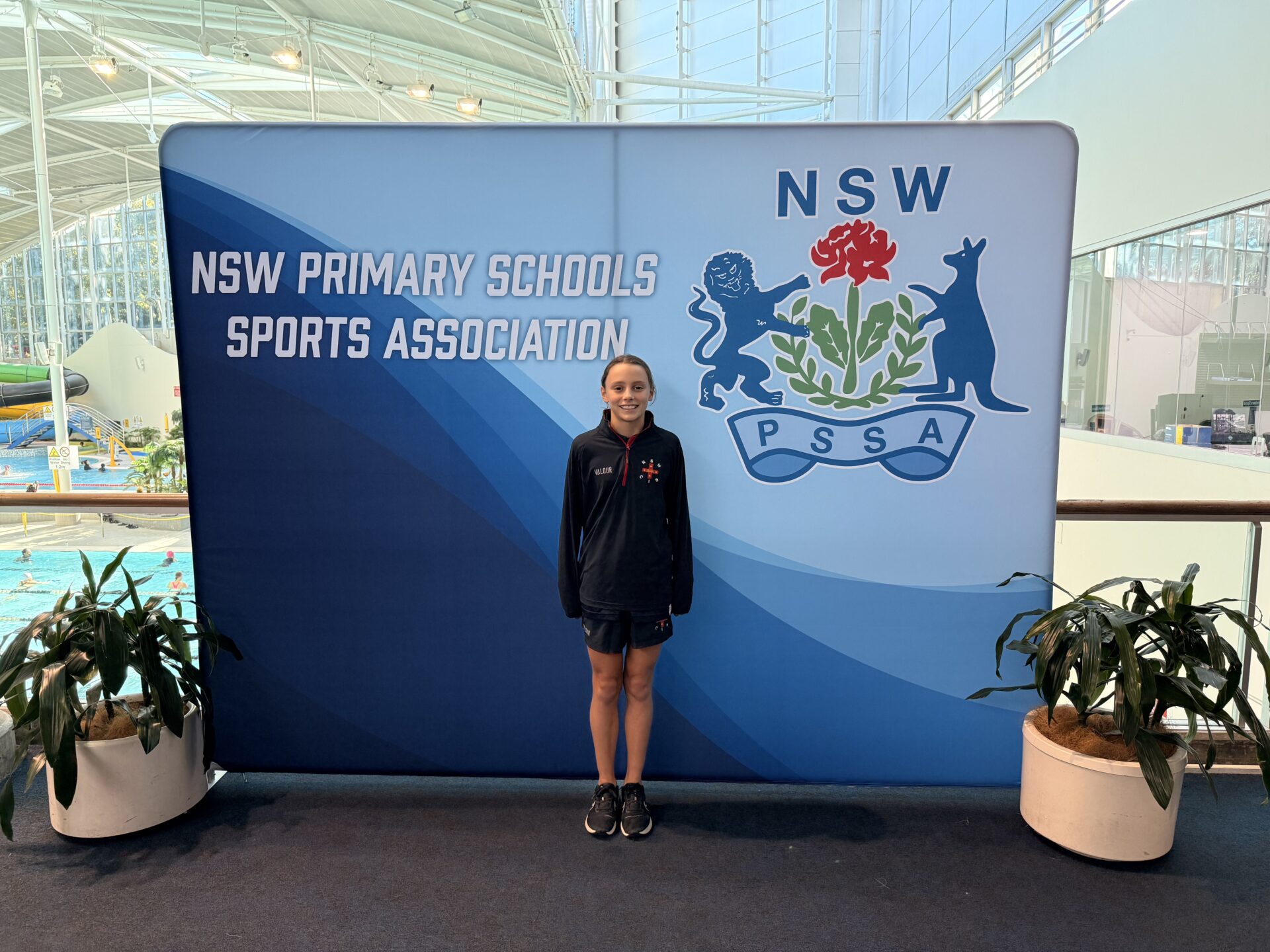 A girl in sportswear stands smiling beside two potted plants and a large NSW Primary Schools Sports Association banner, celebrating her primary sporting success inside a bright indoor swimming pool venue.