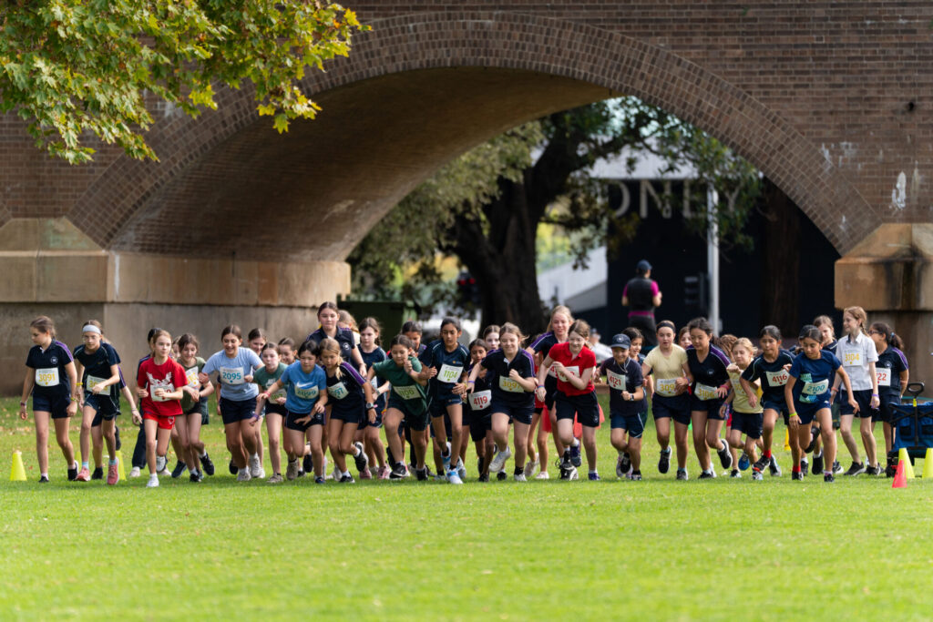 A large group of primary children in sports kit show grit as they start running a race on grass beneath a brick bridge, with trees and cones marking the course.