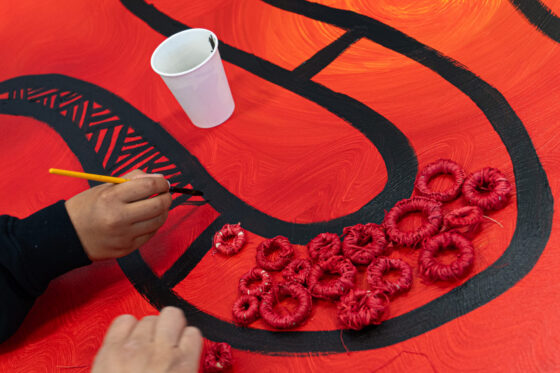 A person’s hand paints a black line with patterns on a red canvas. A white cup and several red, ring-shaped textured objects are also on the canvas.